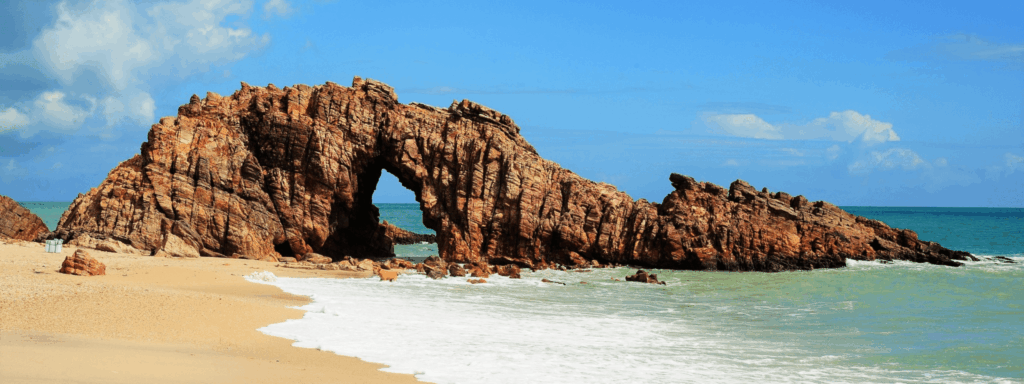 Imagem da Pedra Furada vista de frente, tendo o mar e o horizonte azuis como pano de fundo.