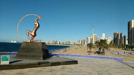 Imagem da Praia de Iracema, com a estátua de Iracema à esquerda, em frente ao mar, e os prédios da orla à direita.